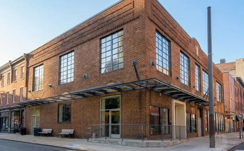 Brick building with large windows, featuring a street-level entrance and benches on a sunny day.