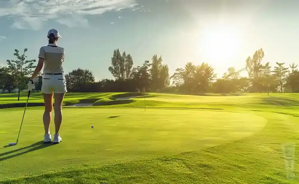 A golfer stands on a green golf course with a golf club, looking towards a bright sun.