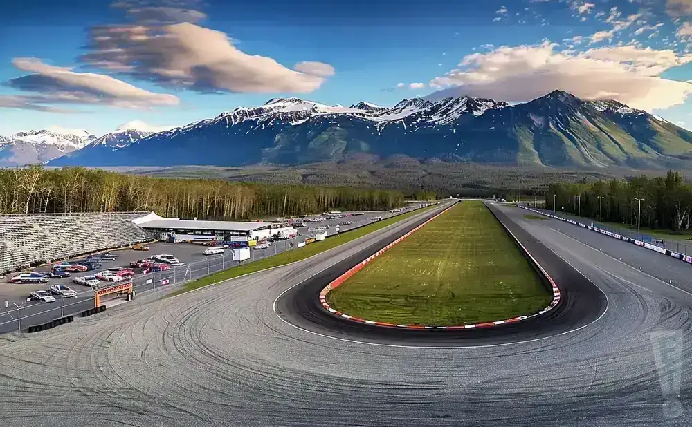 A race track with a grandstand and parking lot, set against a backdrop of snow-capped mountains and trees.