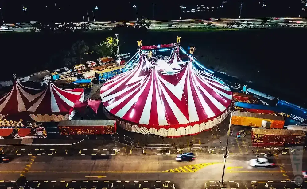 An aerial view at night of a red and white striped circus tent, surrounded by circus trailers and a highway.