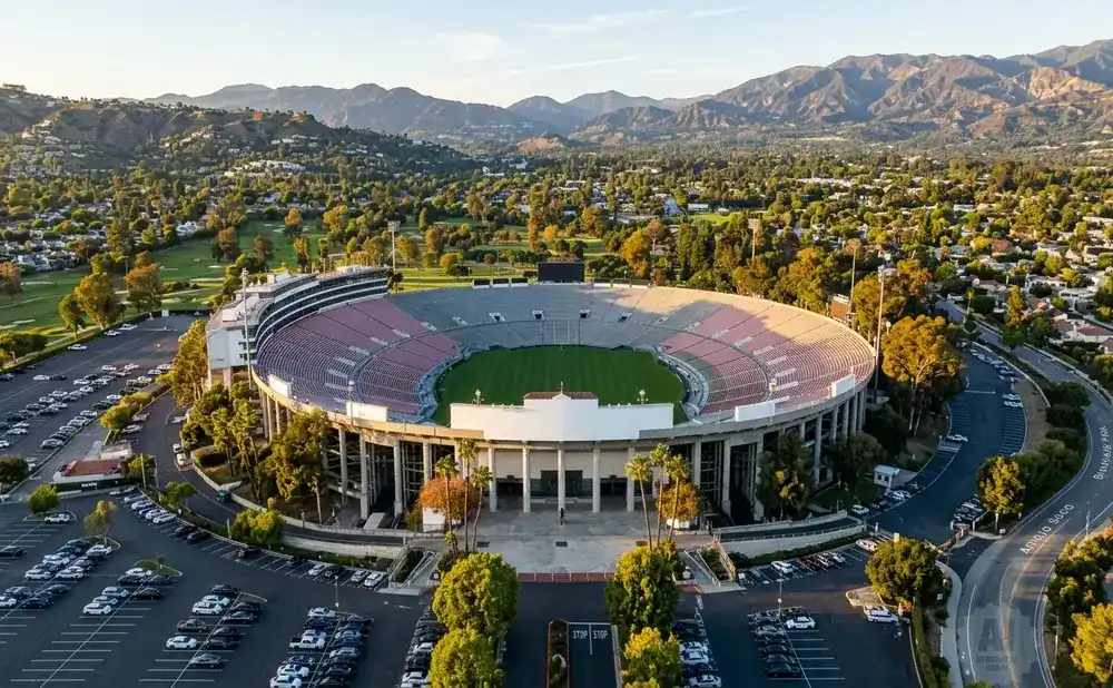 Aerial view of a large, circular stadium with a green field, surrounded by trees and mountains.