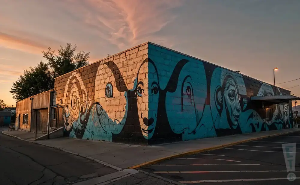 Mural of faces and waves on a brick building at sunset.