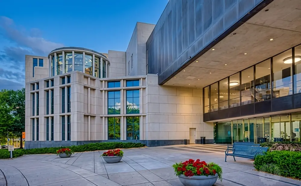 A modern building with a curved glass section and large windows, surrounded by greenery, under a twilight sky.
