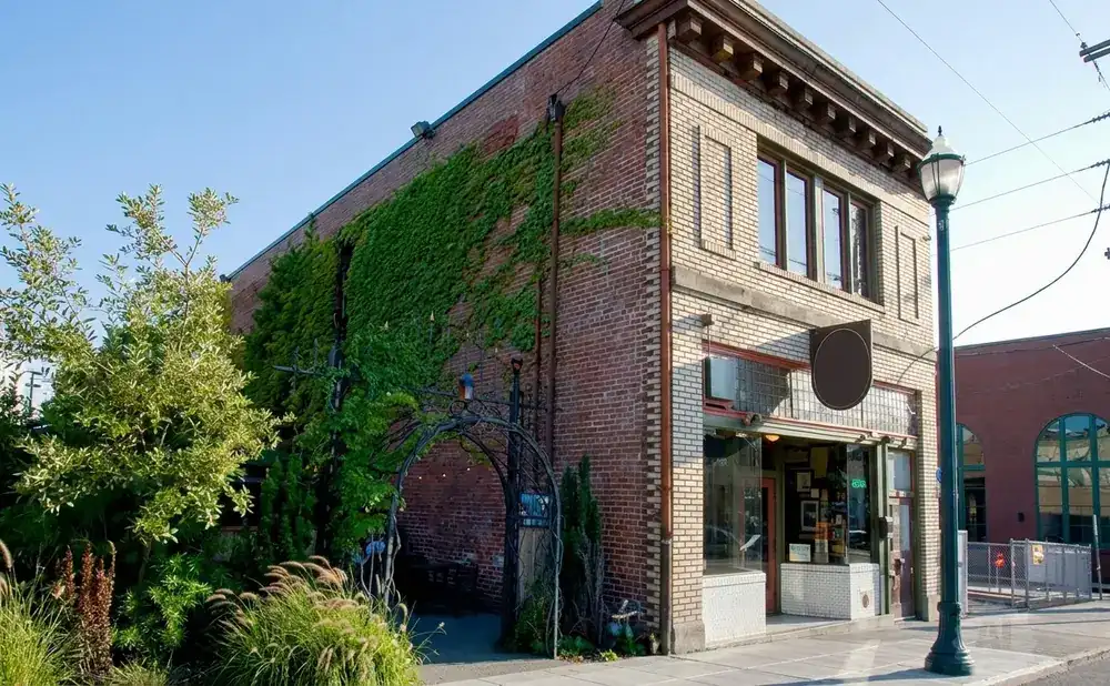 A brick building with ivy, an arched entrance, and a streetlamp stands on a sidewalk under a clear blue sky.