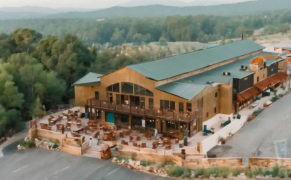 An aerial view of a large, rustic building with a green roof, surrounded by trees and a patio with outdoor seating.