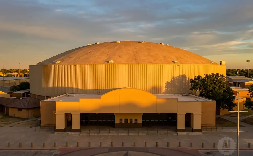 A domed stadium bathed in golden hour light, with a tan facade and a backdrop of trees and sky.