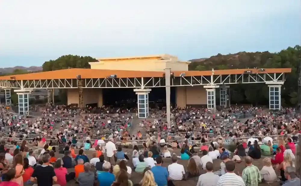 An outdoor amphitheater with a large audience seated on tiered rows facing a stage structure.