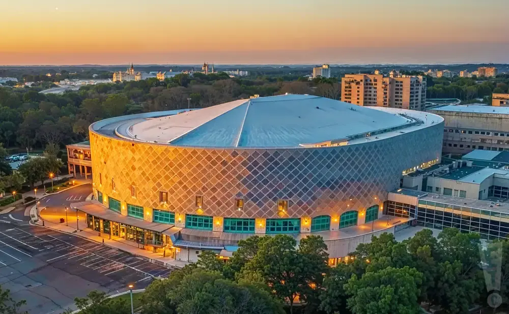 Aerial view of a circular arena with a patterned facade and a large roof, set against a sunset sky.