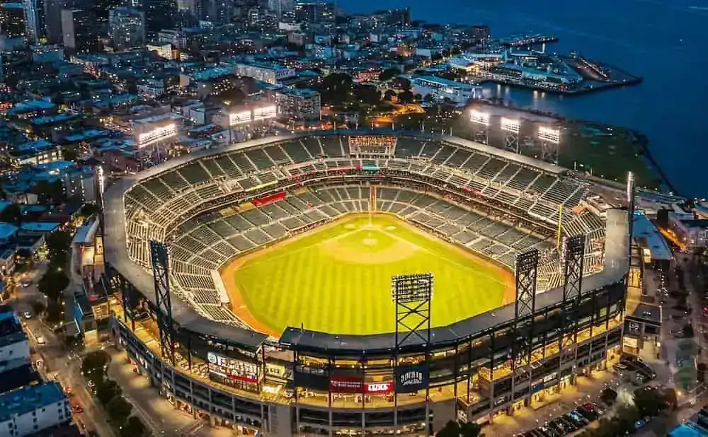 Aerial view of a baseball stadium at dusk, with the city lights and bay in the background.