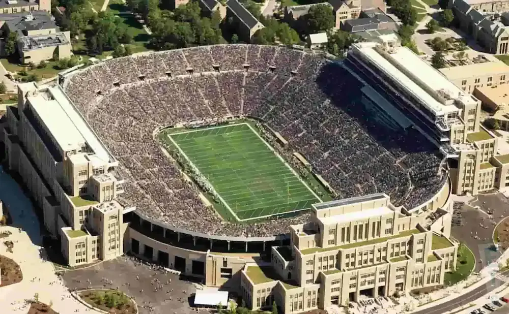 Aerial view of a packed football stadium with a green field, surrounded by stone buildings and trees.