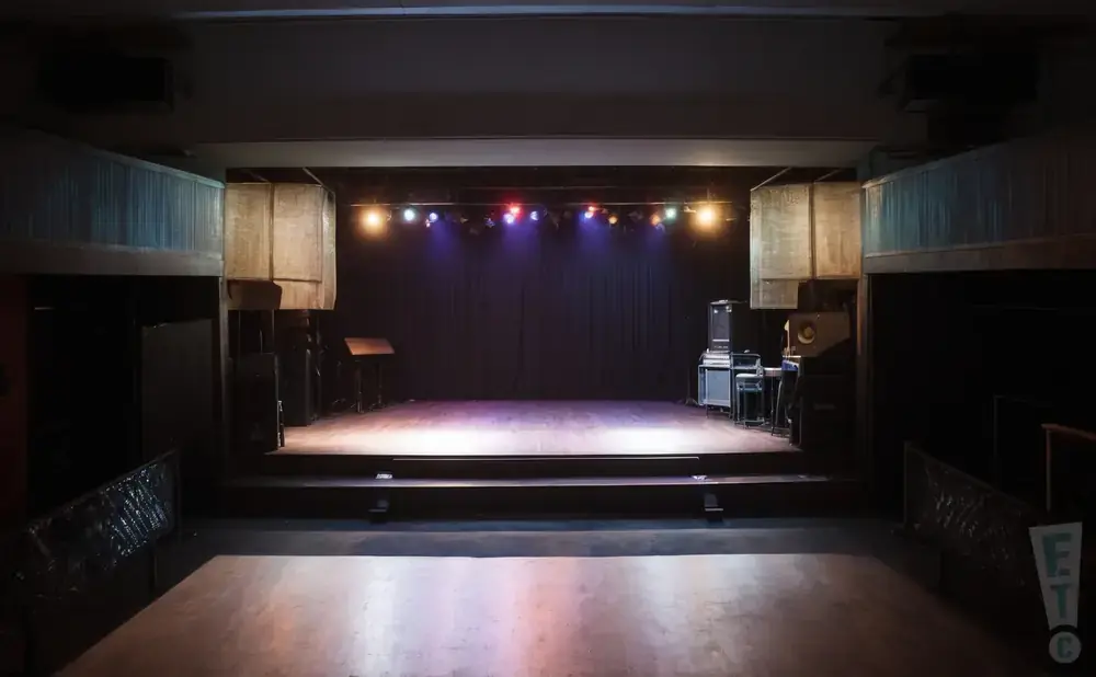 Empty concert hall stage with lighting rig, sound equipment, and a black curtain backdrop.