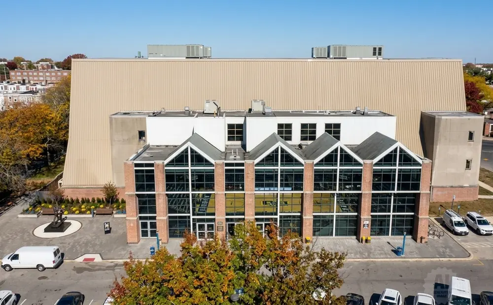 Aerial view of a university athletic facility with large glass windows, brick accents, and a tan metal exterior, surrounded by trees and parking lots.