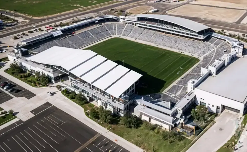 Aerial view of a stadium with a green field, tiered seating, and white-roofed buildings.