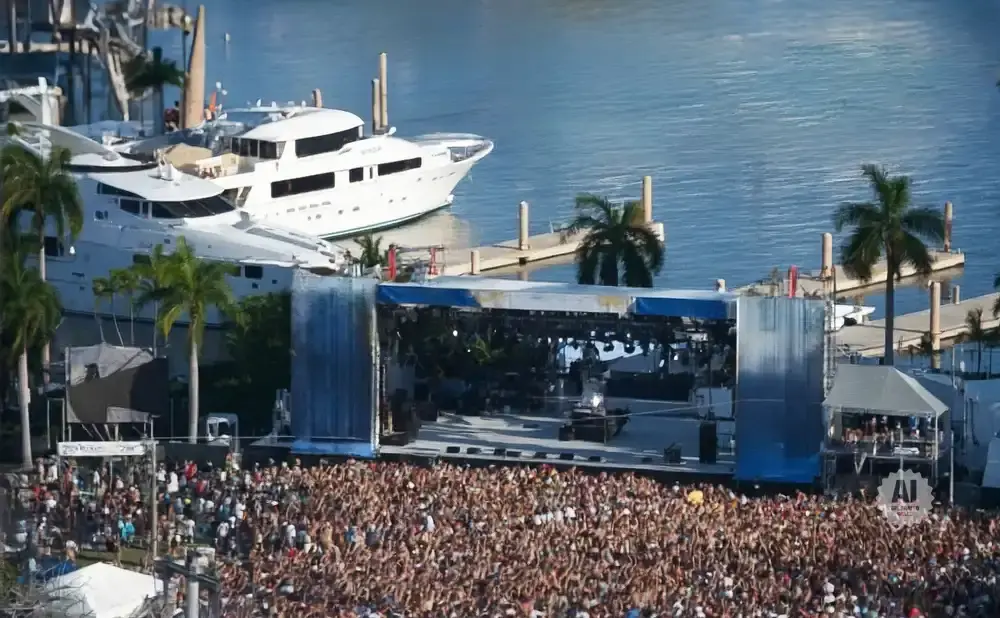 Large outdoor concert crowd in front of a stage with yachts docked in the background.