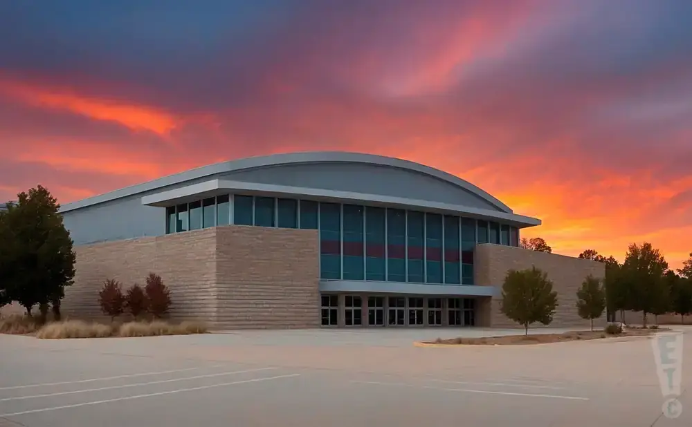 A modern building with a curved roof and large windows stands under a vibrant orange and red sunset sky.