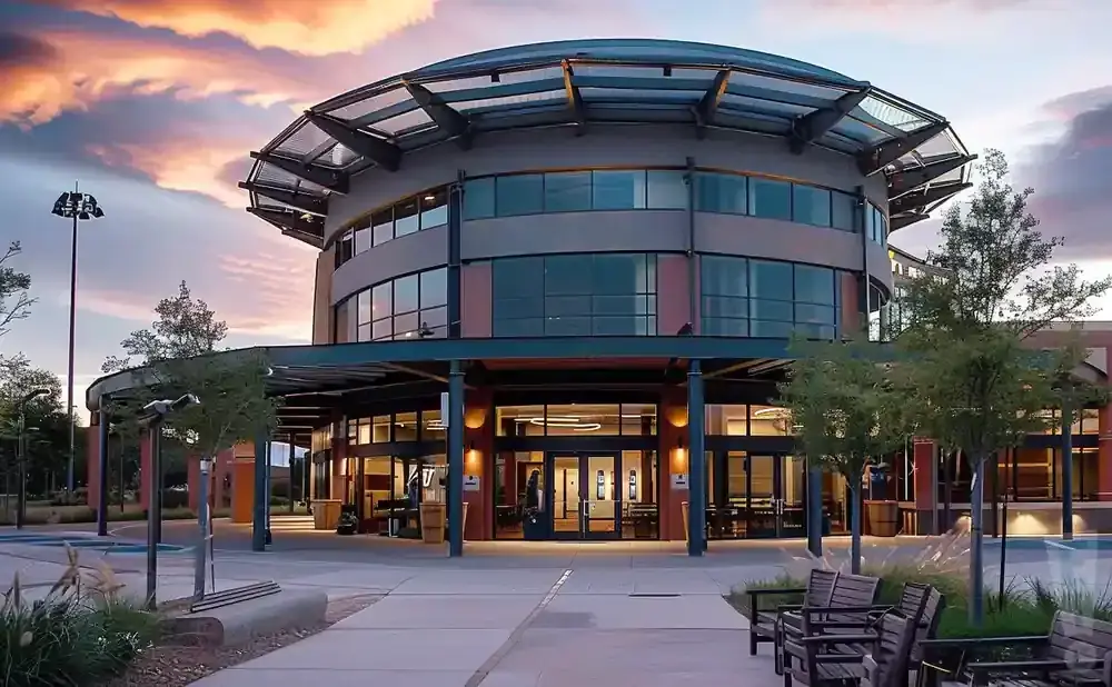 Modern circular building with a glass roof and large windows, with trees and benches in front during sunset.