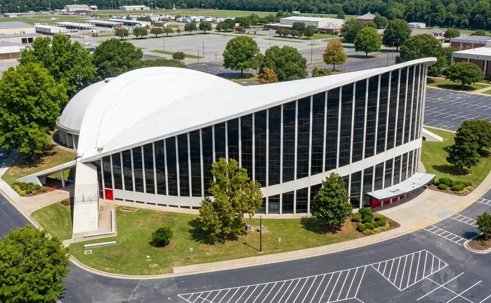 Aerial view of a modern building with a curved white roof and large glass windows, surrounded by trees and parking lots.