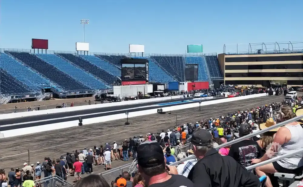 Spectators at a drag race in a stadium, with blue bleachers and a race track visible under a clear blue sky.
