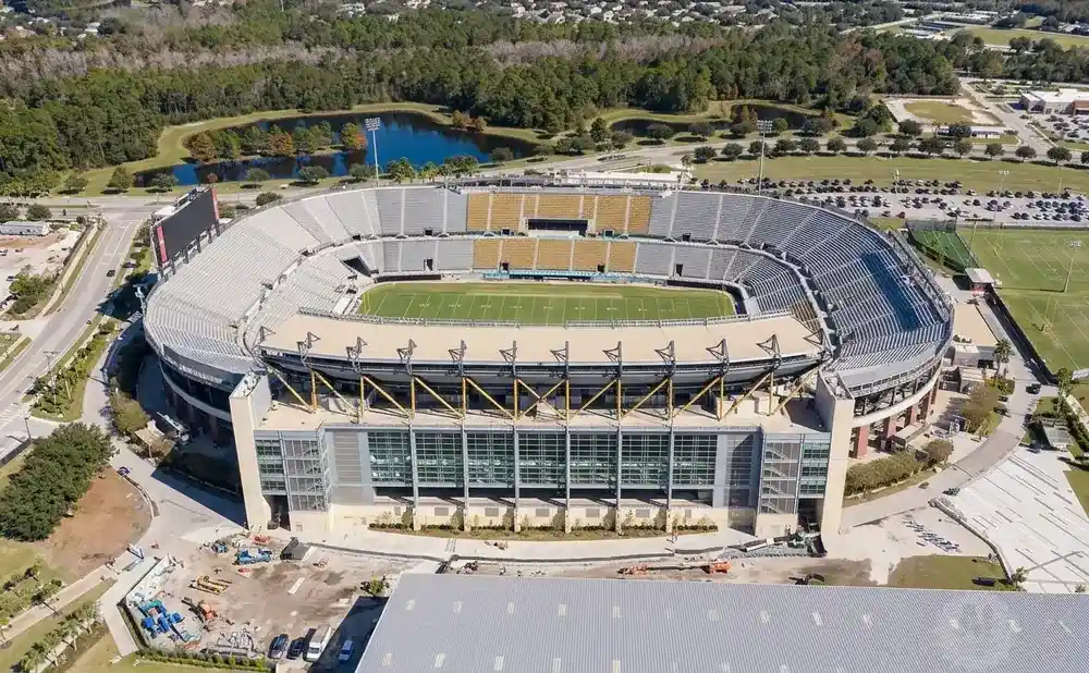 Aerial view of a football stadium with a large scoreboard, surrounded by trees and practice fields.