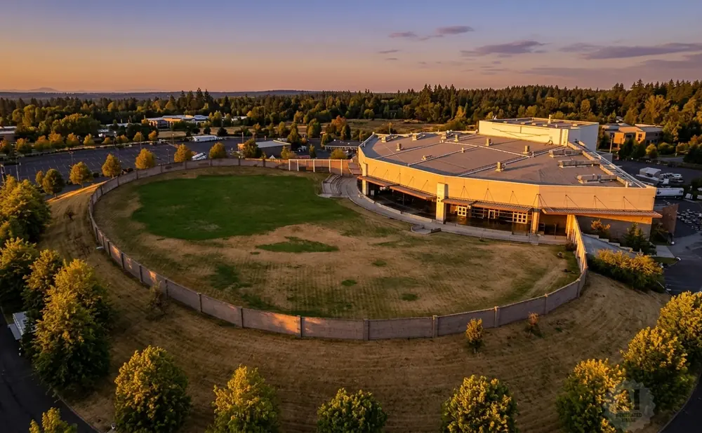 Aerial view of a large building with a rounded facade, surrounded by a grassy field and trees at sunset.