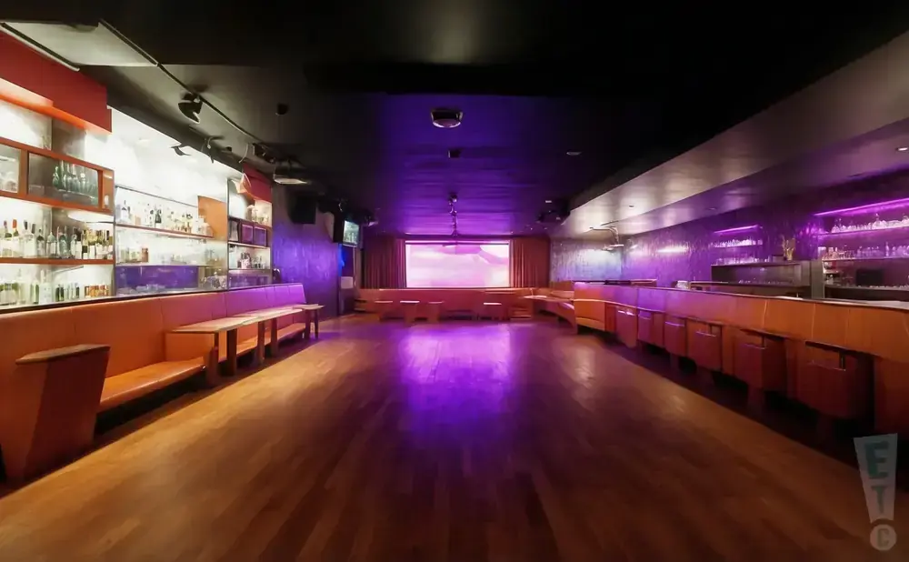 An empty, dimly lit nightclub with orange booths, a bar, and a large projection screen.