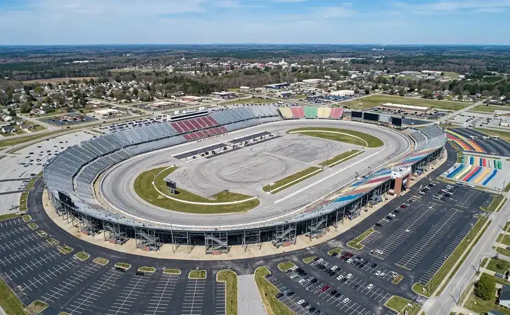 Aerial view of a large oval race track with grandstands and surrounding parking lots and buildings.