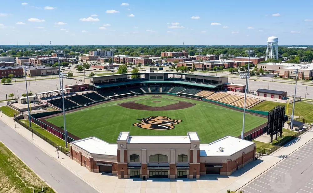 Aerial view of a baseball stadium with green field, stands, and surrounding buildings under a blue sky with clouds.