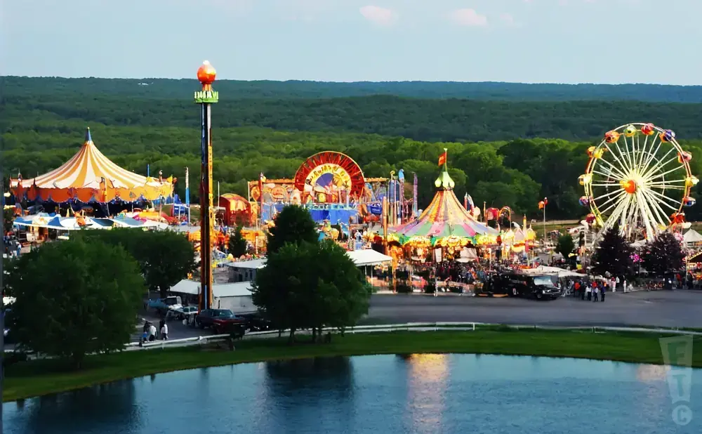 A vibrant amusement park with a Ferris wheel, carousel, and carnival rides, set against a backdrop of green trees and a calm lake.