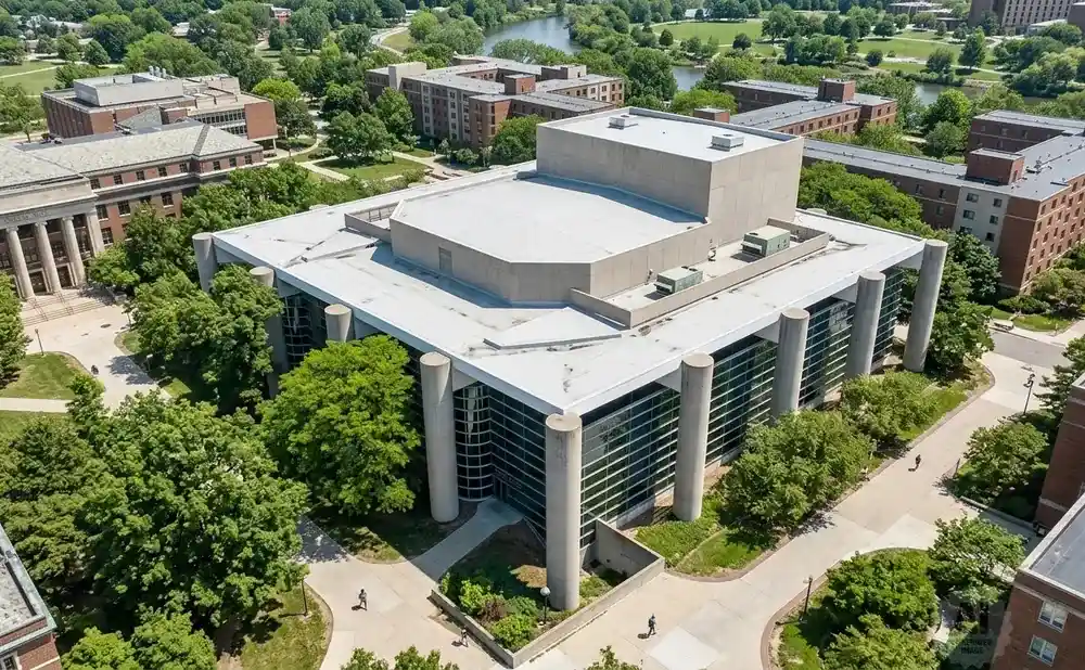 Aerial view of a modern university building with large glass windows, concrete pillars, and a flat roof, surrounded by trees and other campus buildings.