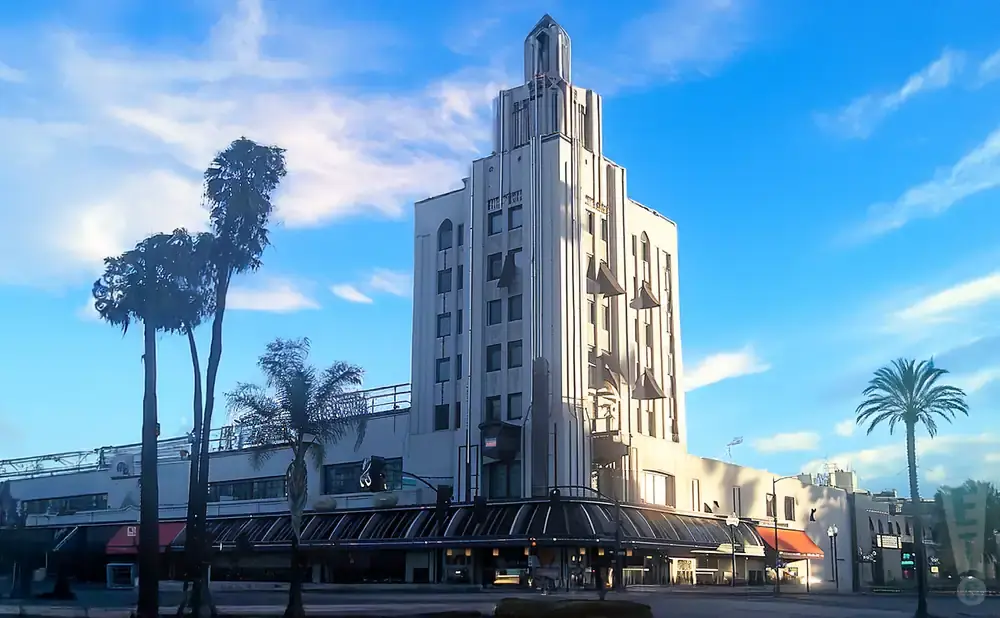 Art Deco building with a tiered tower and awning-covered shops, framed by palm trees under a bright blue sky.