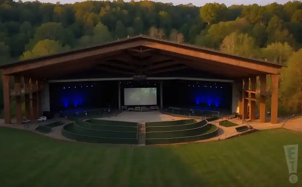 Outdoor amphitheater with a covered stage, screen, and tiered seating surrounded by grass.