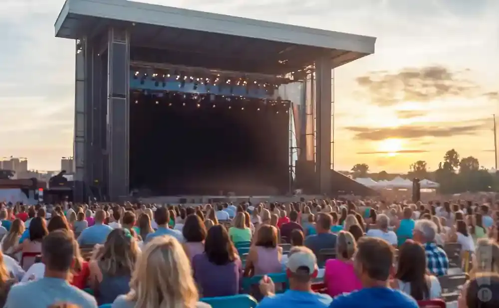 A large outdoor concert crowd watches a band play on a stage at sunset.