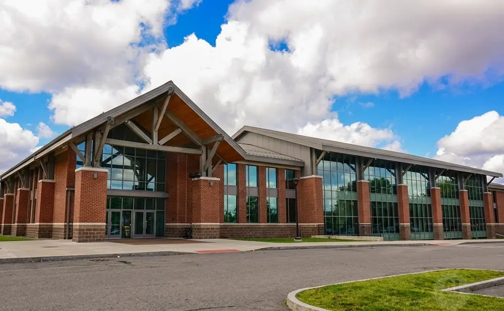 Modern brick building with large glass windows and a wooden-beamed pitched roof against a cloudy blue sky.
