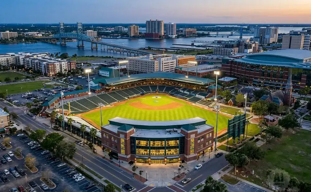 Aerial view of a baseball stadium at dusk with city skyline and a bridge in the background.