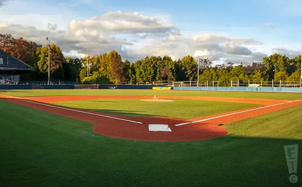 A baseball field with manicured grass and clay baselines under a partly cloudy sky.