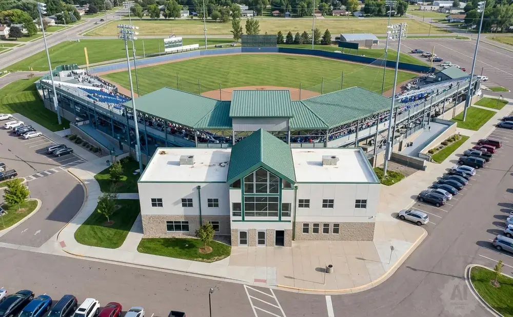 Aerial view of a baseball stadium with a main building, bleachers, and a parking lot filled with cars.