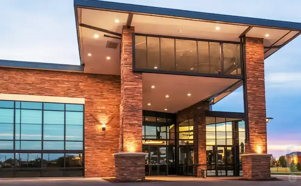 Modern building entrance with stone facade, large glass windows, and an overhang lit by recessed lights.