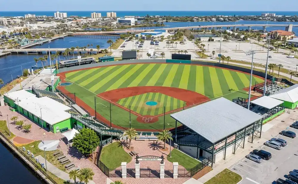 Aerial view of a baseball field with green and red infield, surrounded by buildings, a parking lot, and a body of water.