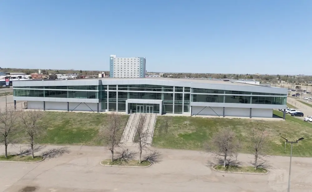 Modern glass-fronted office building with stairs leading to the entrance, surrounded by trees and parking lots.