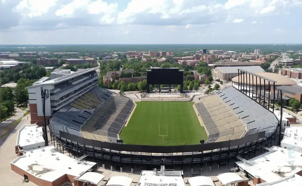 Aerial view of an empty football stadium with a large scoreboard, green field, and surrounding buildings.