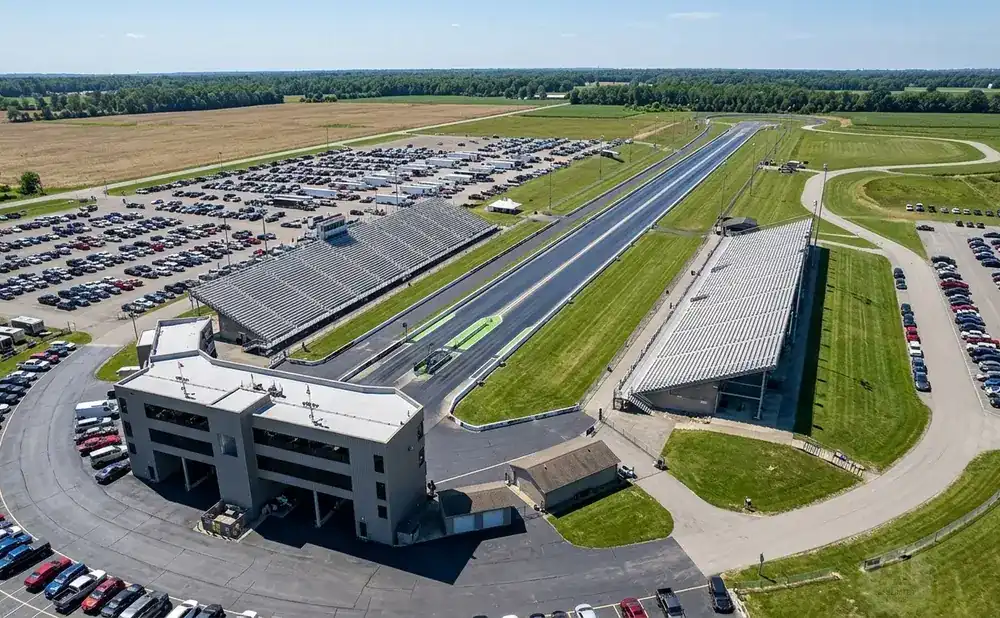 Aerial view of a drag strip with grandstands and large parking lots filled with cars.