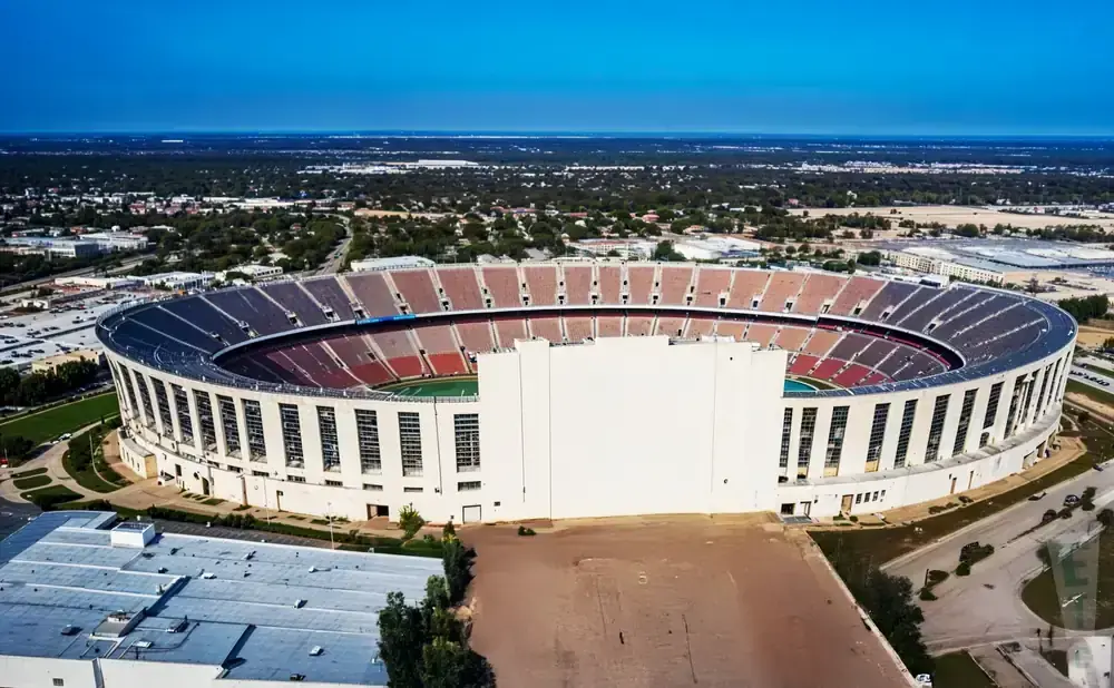 Aerial view of a large, oval stadium with blue and red seats, set against a bright blue sky.