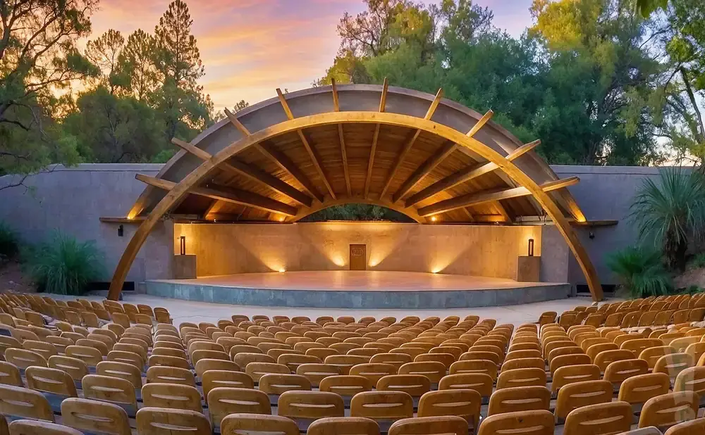 Outdoor amphitheater stage with rows of wooden seats under a curved wooden canopy, lit by warm lights.