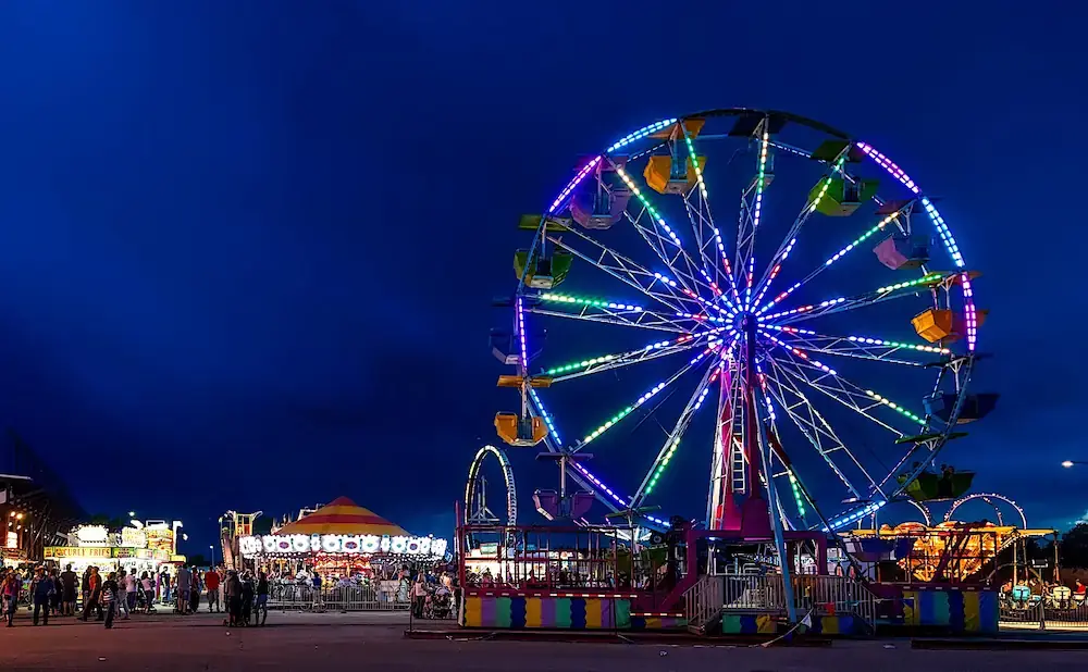 A brightly lit Ferris wheel and carnival rides glow against a deep blue night sky at a fair.