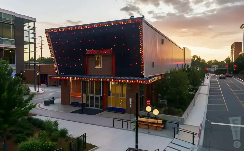 Modern building with red and gold lights on the roof, at dusk with a street and cars.
