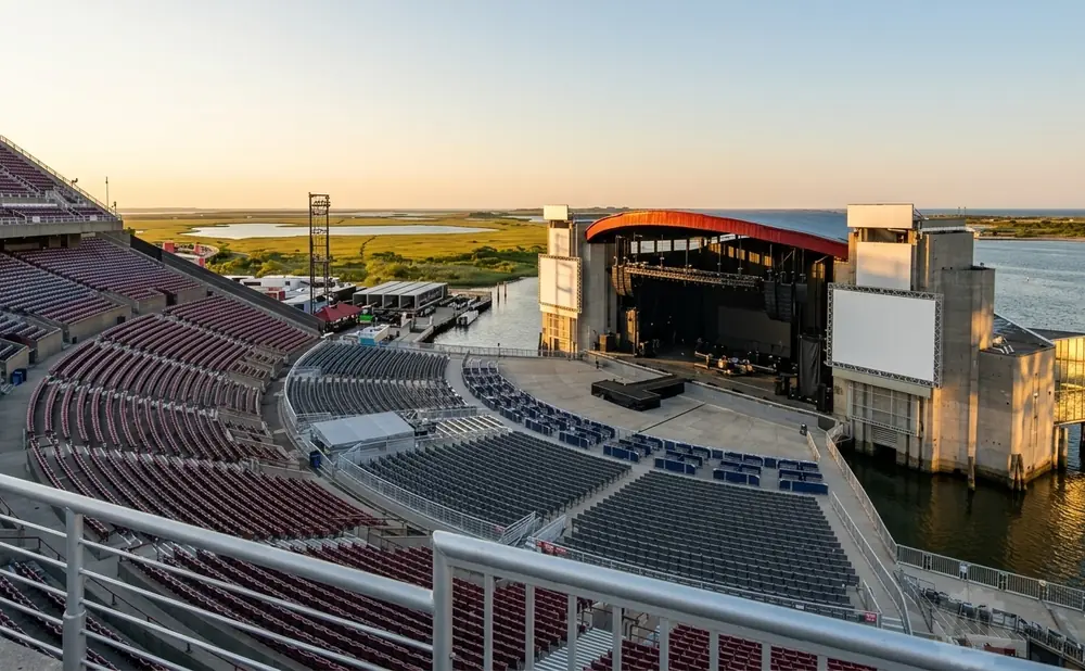 Outdoor amphitheater with a stage on a body of water, set against a marshy landscape and clear sky at sunset.