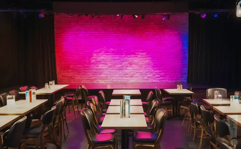Empty comedy club with a brick stage lit pink and blue, and tables and chairs in the foreground.