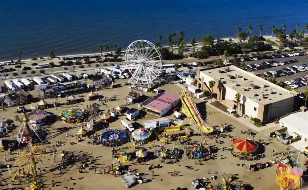 Aerial view of a busy seaside fair with rides, attractions, and a parking lot.