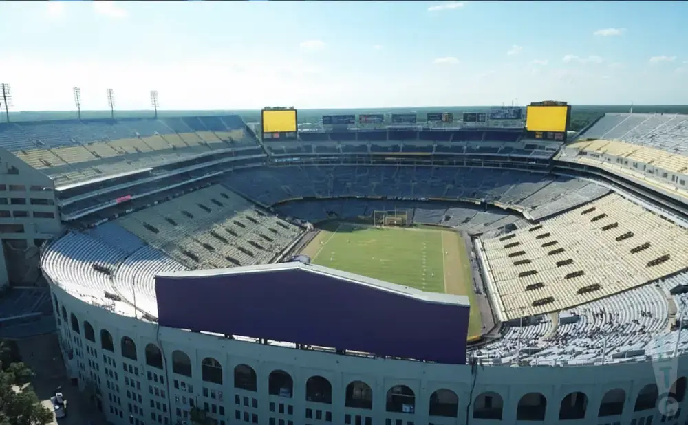 Aerial view of a large, empty football stadium with yellow and blue seats, a green field, and two large scoreboards.