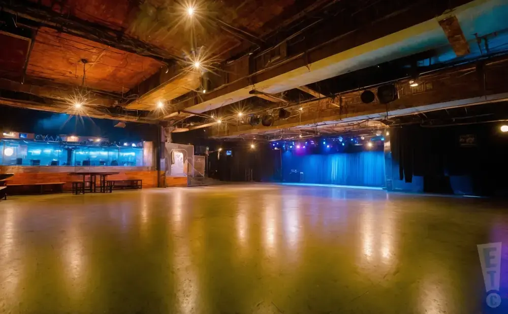 An empty, industrial concert venue with a stage, blue curtains, and a polished concrete floor reflecting stage lights.